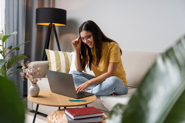 Smiling woman sitting cross legged on a beige sofa working on a laptop in a modern living room